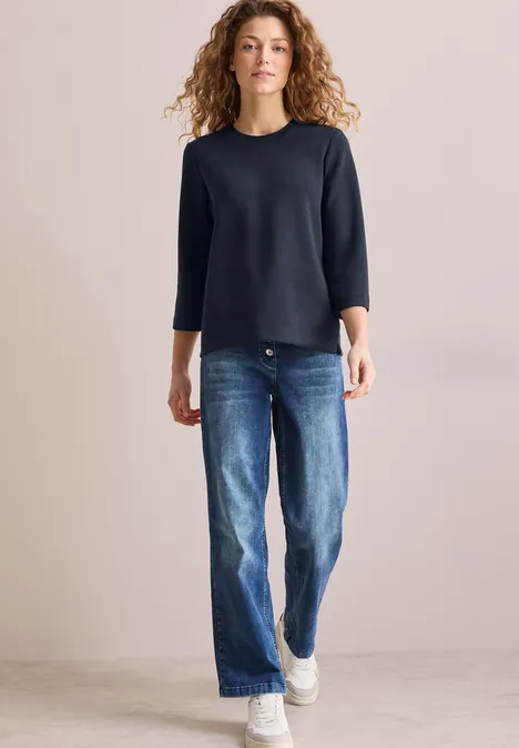 Frau mit lockigen Haaren trägt dunkles Shirt, Jeans und weiße Sneakers.